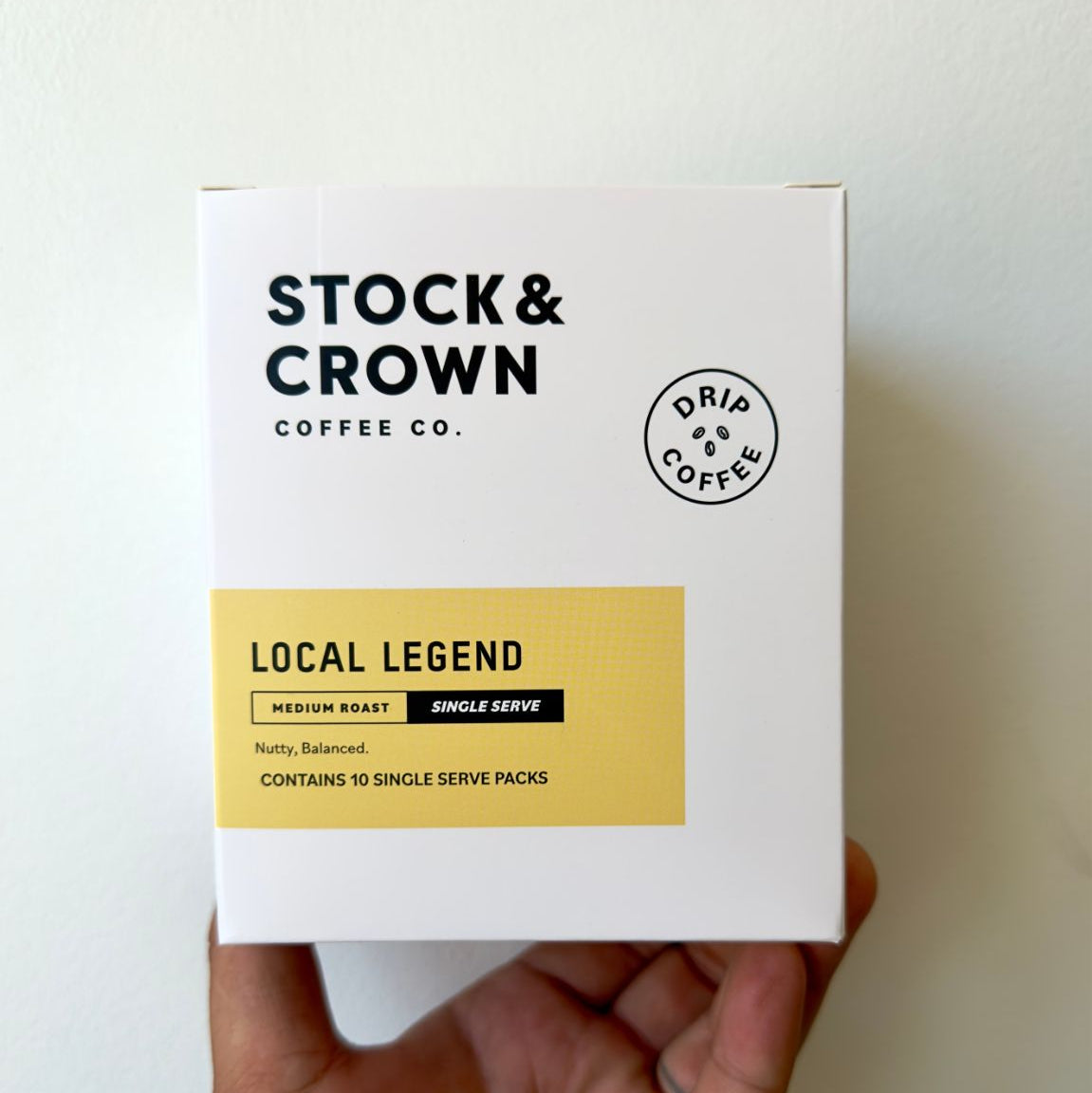 A hand holding a white box of Stock & Crown Coffee Co. Local Legend single-serve coffee packs against a plain off-white wall.